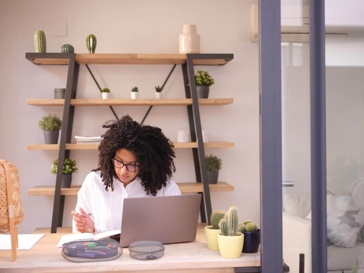 A Black professional woman seated at a desk focuses on her laptop and notebook.