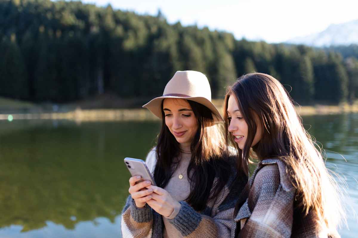 Two female friends look at smartphone by lake at sunset