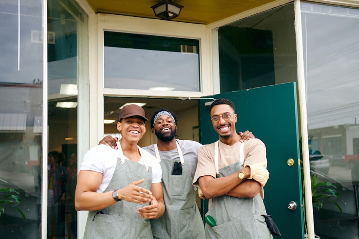 A trio of Black men wearing work aprons laughs together outside of a store front.