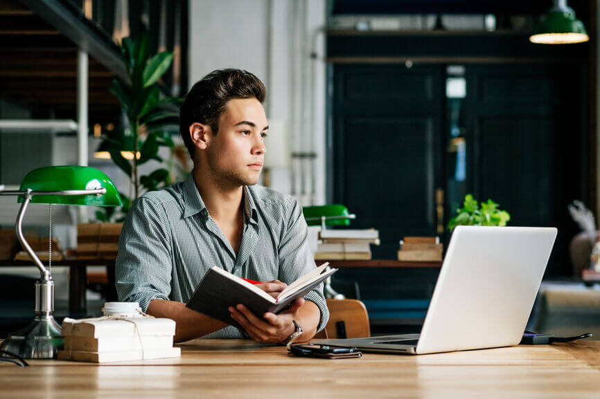 Young man writing in a journal and working on a laptop in the library.