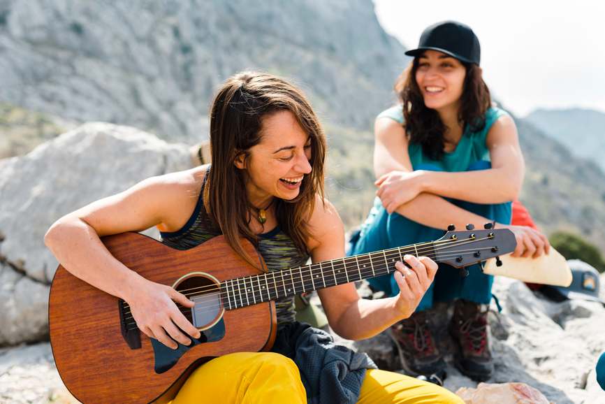 Female camper plays acoustic guitar for female friend in mountains