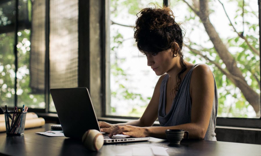 Woman sitting by large windows working on her laptop at a dark brown desk