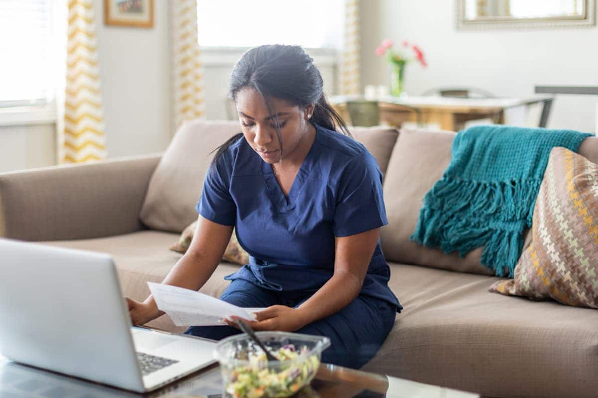 A woman in scrubs seated on a couch works at a laptop.