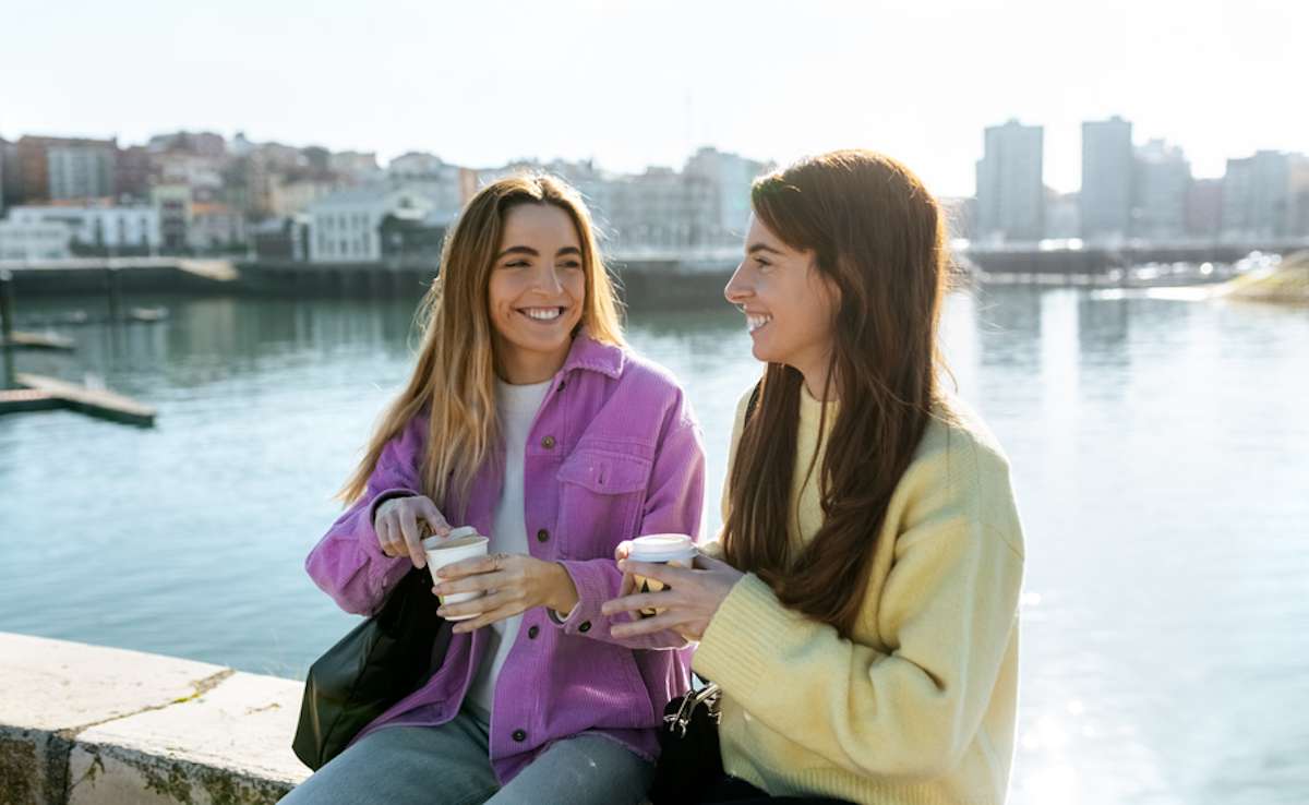 Two young women pause outside in a city to drink coffee