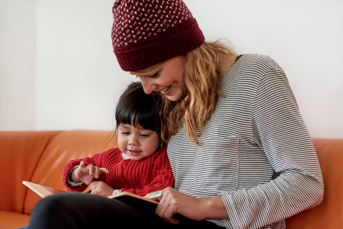 Young mom in stocking cap reads to daughter on orange couch