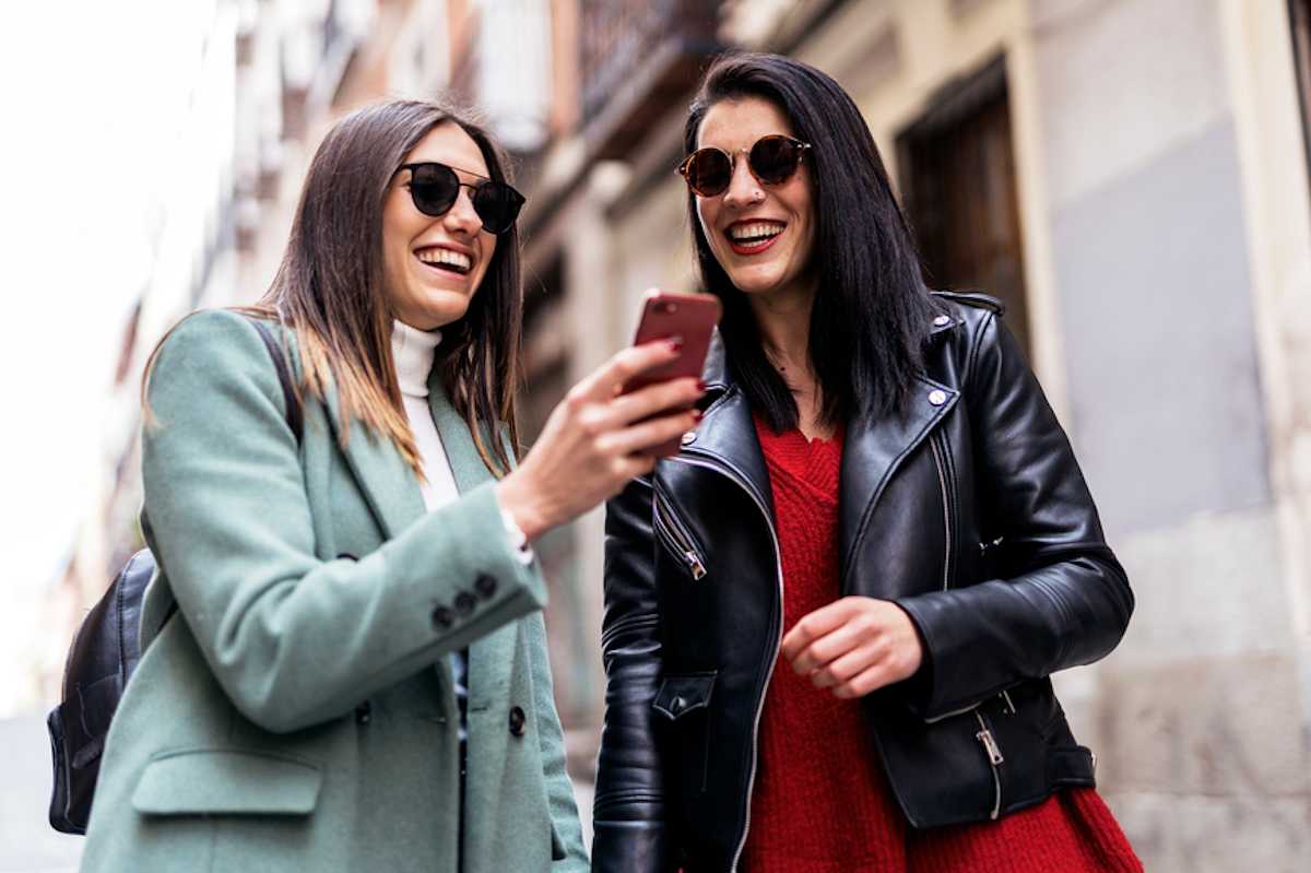 Two young women outdoors in a city share a laugh at something on a smartphone