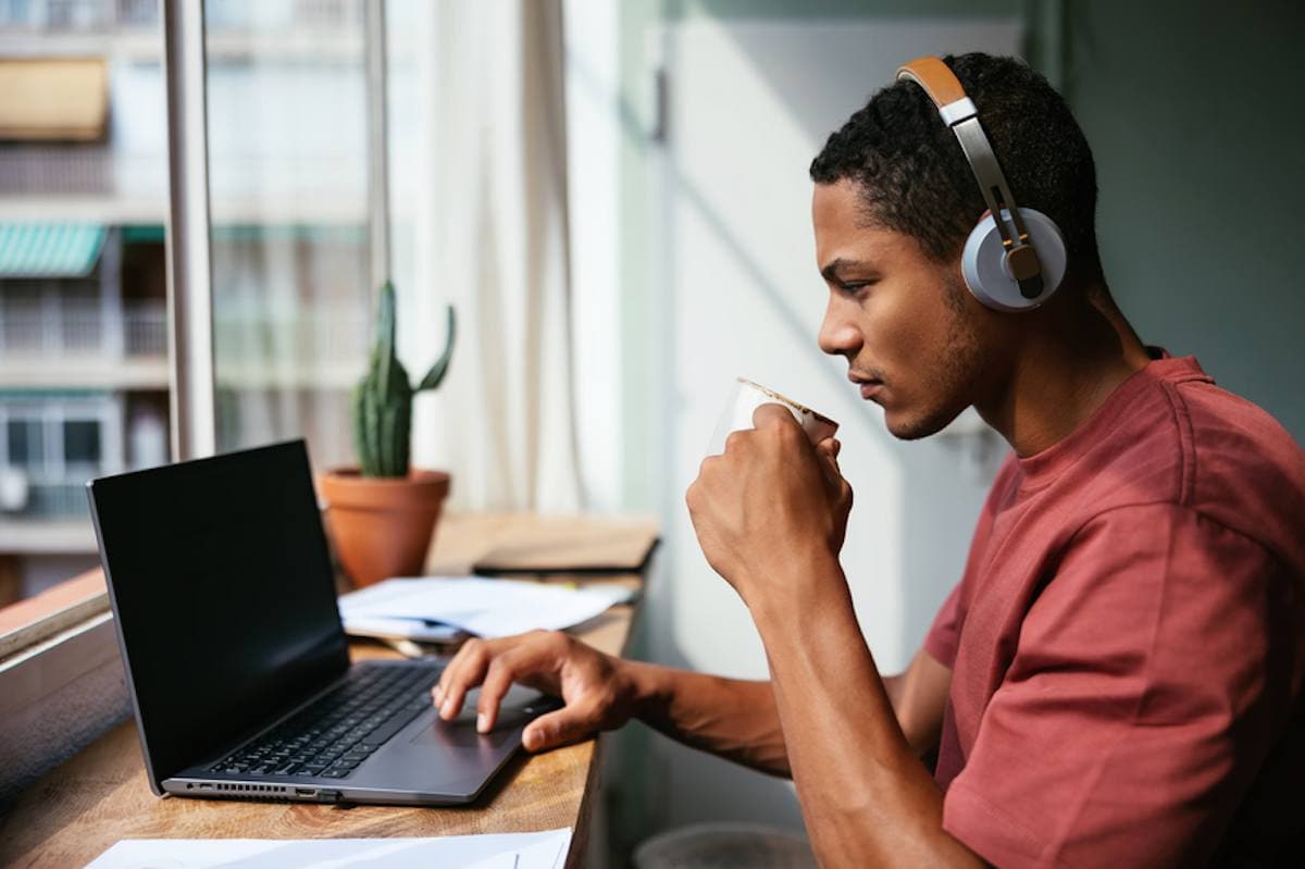 Un joven serio tomando una bebida caliente y escuchando música mientras trabaja en casa