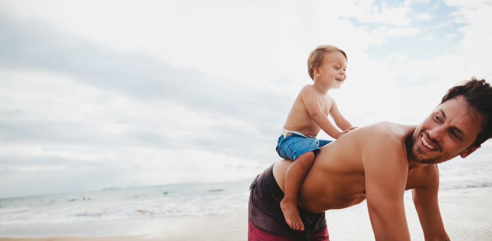 Toddler rides on his father's back while playing on the beach. 