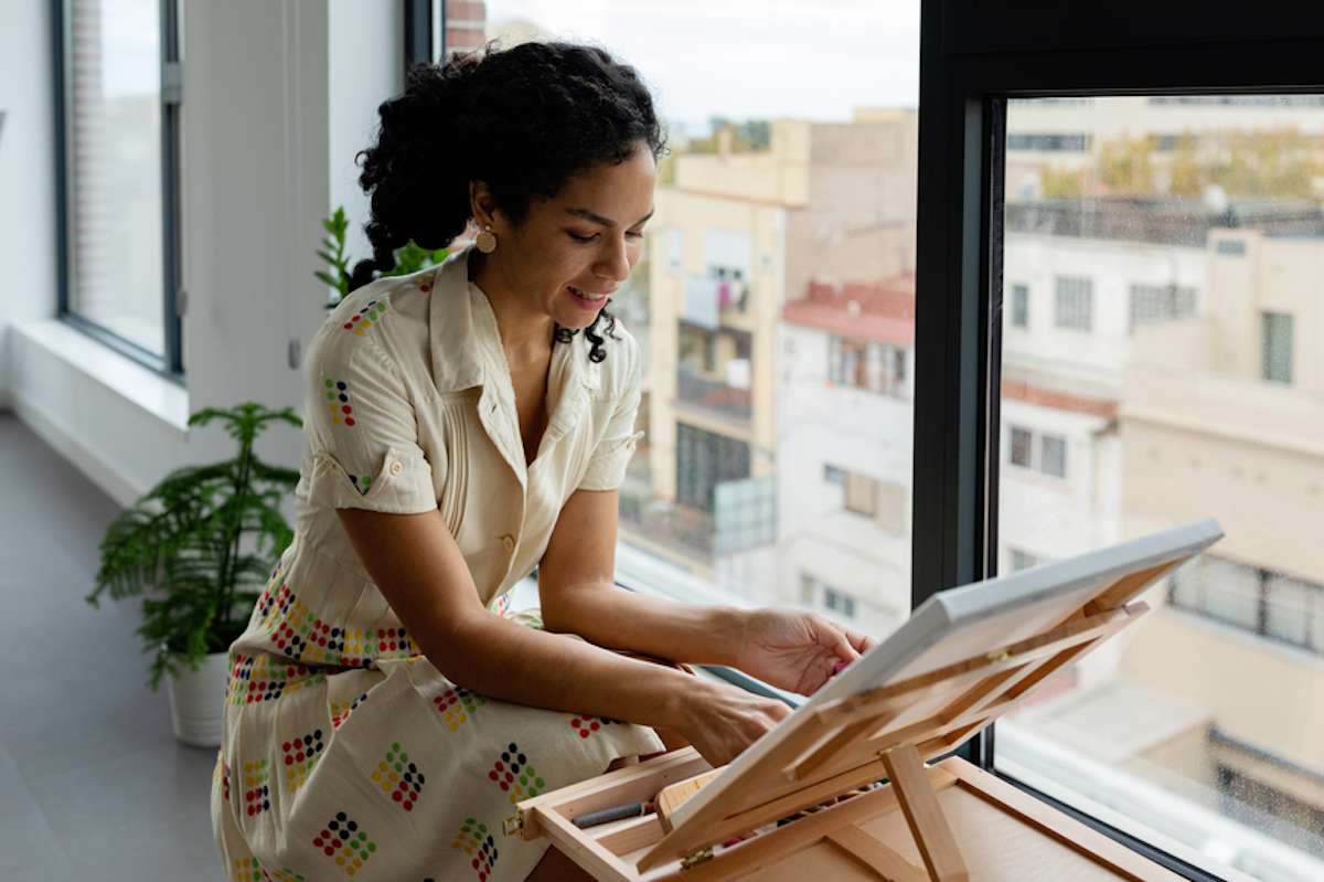 Una mujer parada frente a una ventana pinta sobre un lienzo.