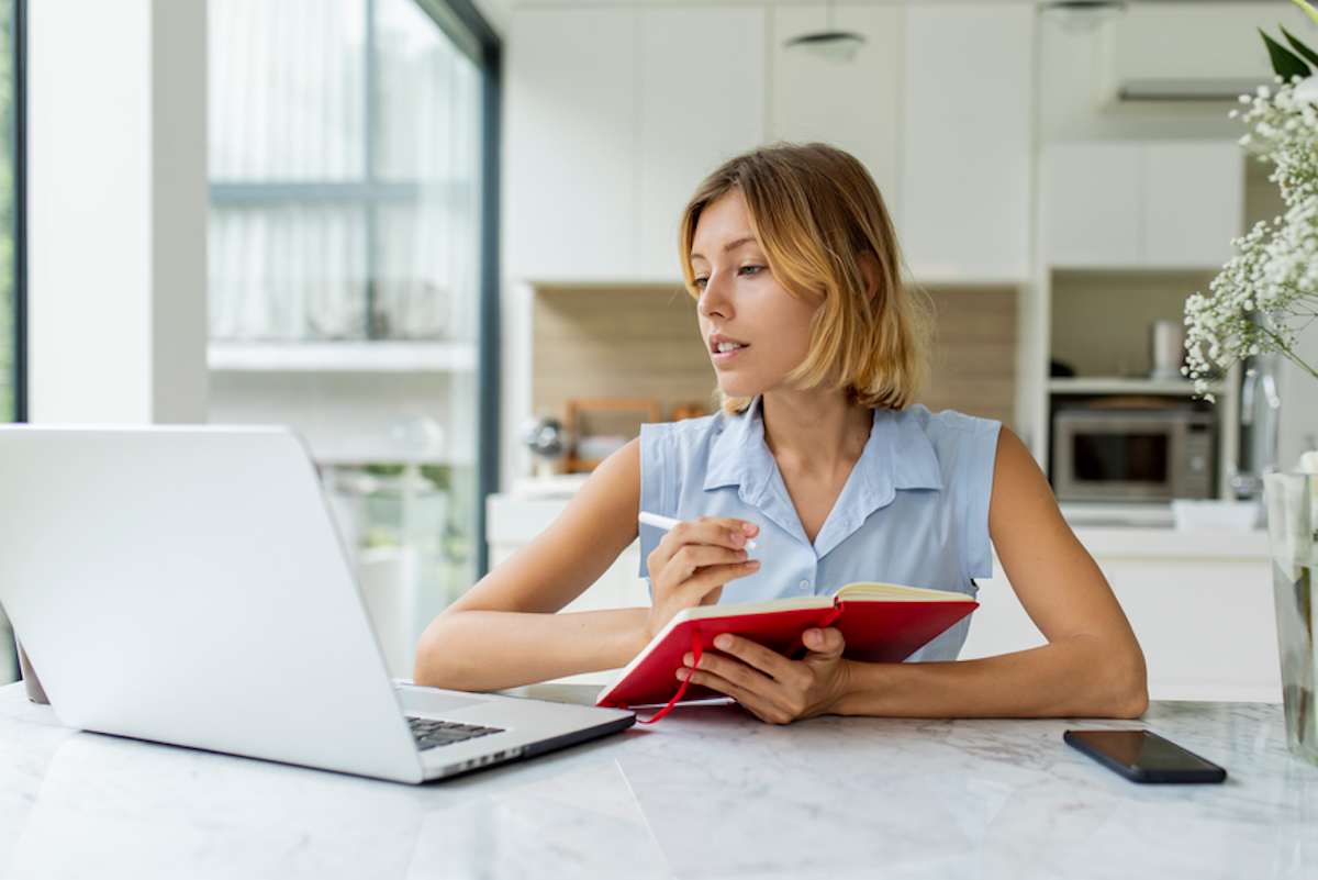 Young woman working from home holds a notebook and pen and looks at a laptop computer