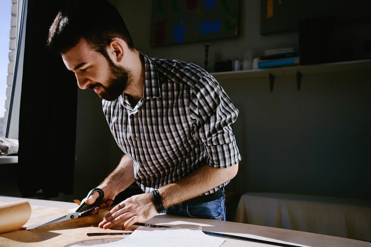 Custom clothier in checkered shirt with large scissors cutting a clothing pattern