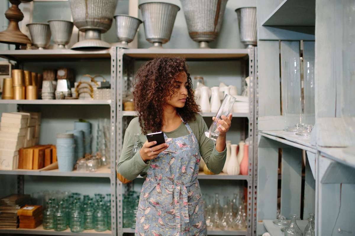 Young woman in print apron inspects glass vase in storeroom