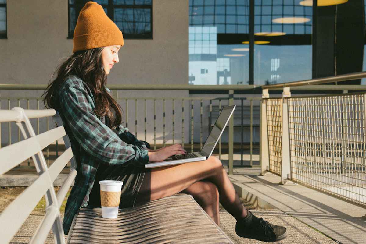 Una mujer joven sentada en un banco del parque trabaja en su computadora portátil.