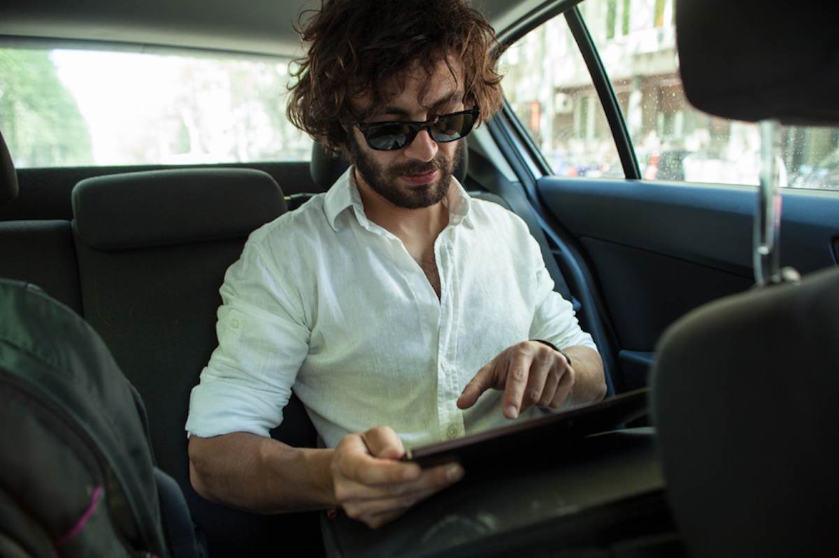 一个穿着随意的年轻人坐在汽车后座上用平板电脑工作