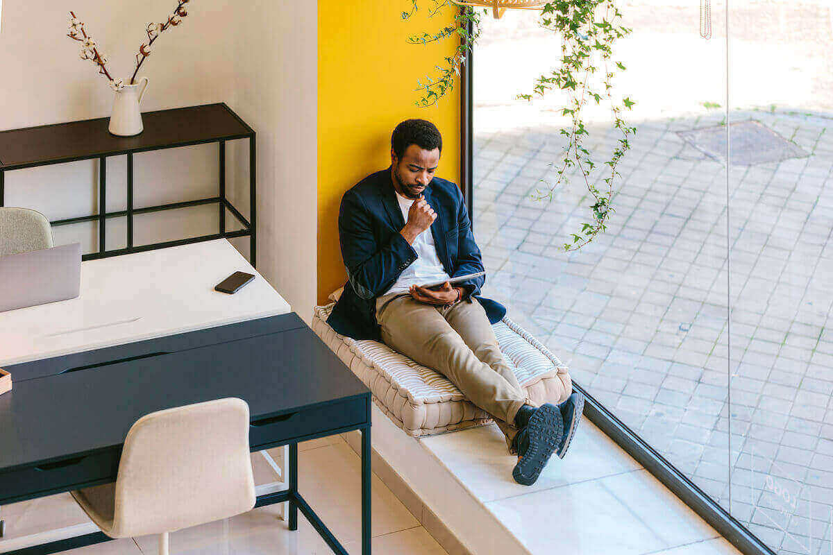 A Black man seated in a large windowsill looks at a tablet. 