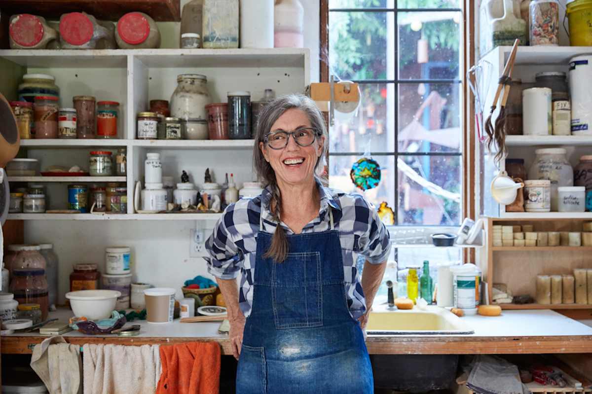 A smiling mature woman in overalls stands in her pottery studio