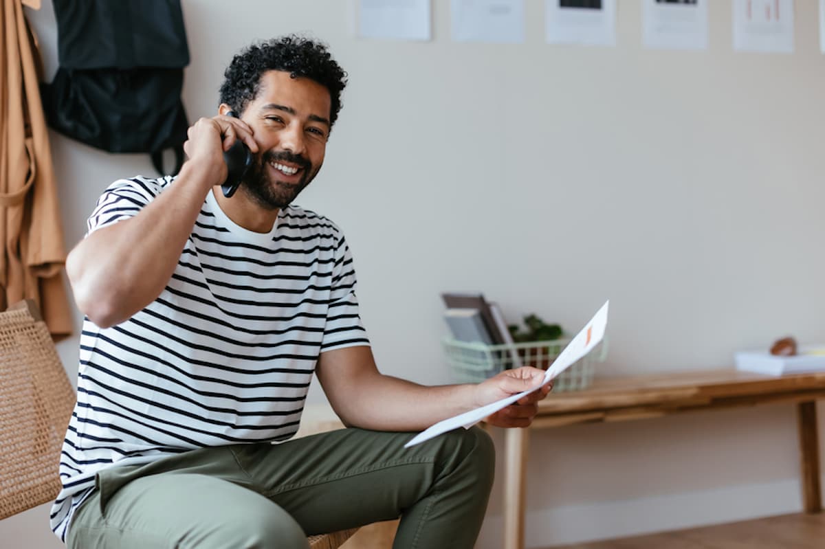 A Black man smiles as he talks on the phone and holds papers.