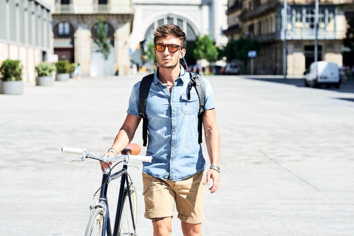 Man in denim shirt wearing sunglasses walks his bicycle down sunny street