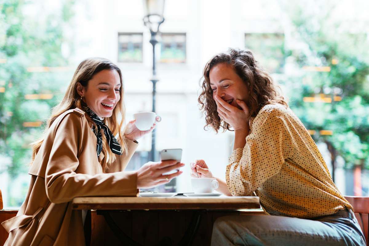 Two stylish women at a coffee shop laugh at something on a smartphone