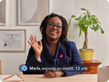 Maria, une experte en impôts avec 12 ans d’expérience, souriante et saluant de la main depuis son bureau à domicile.