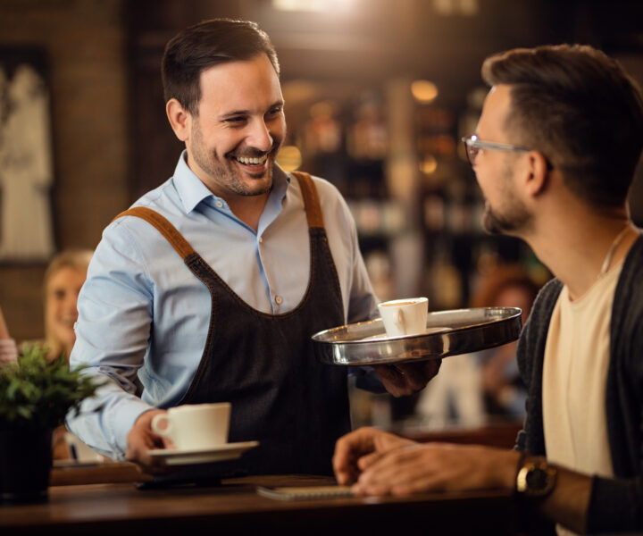 Happy waiter talking to a man while serving him coffee in a cafe.