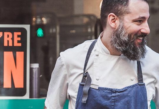 A person in a white shirt and apron stands near a table.