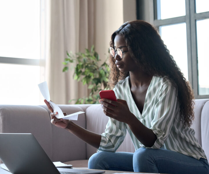 Young black woman holding paper paying bills in mobile application on cellphone.