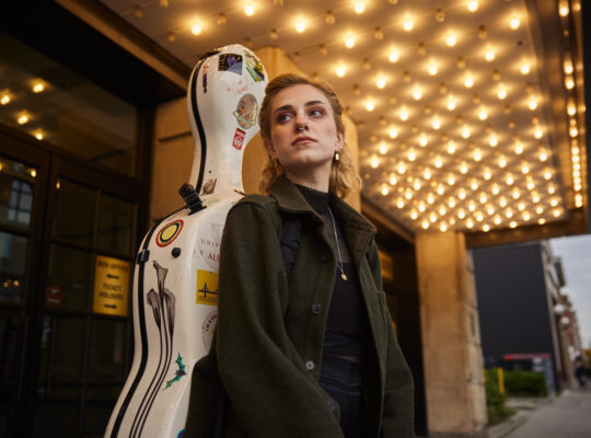 A person stands in a hall with a guitar on her back.