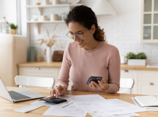 A woman is sitting at a table with a laptop, calculator, and cell phone.