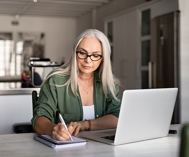 A woman writing on a notebook with a laptop on the table