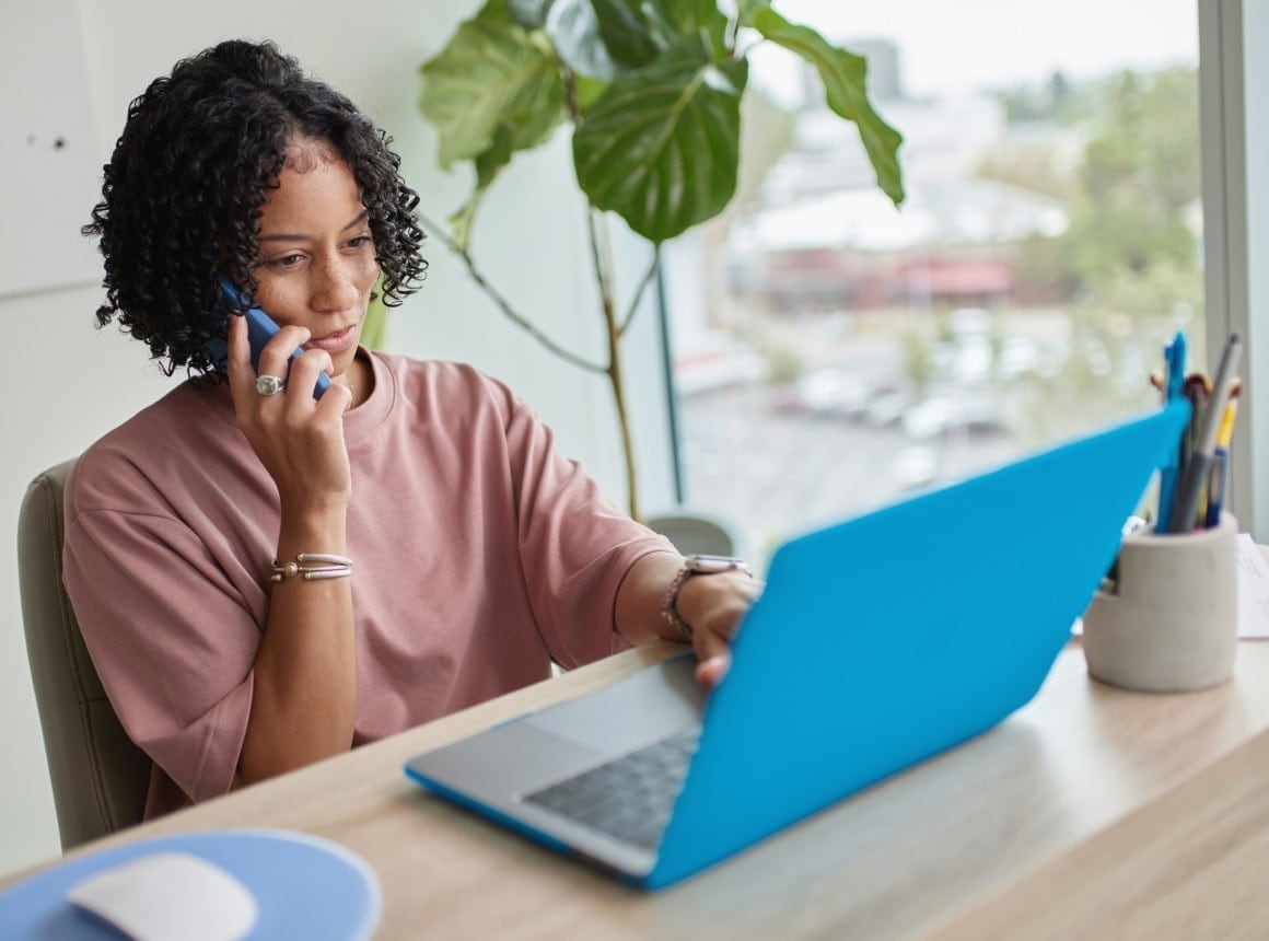 A woman is sitting down at her desk with a laptop. She has a phone to her ear and is looking at her computer screen.