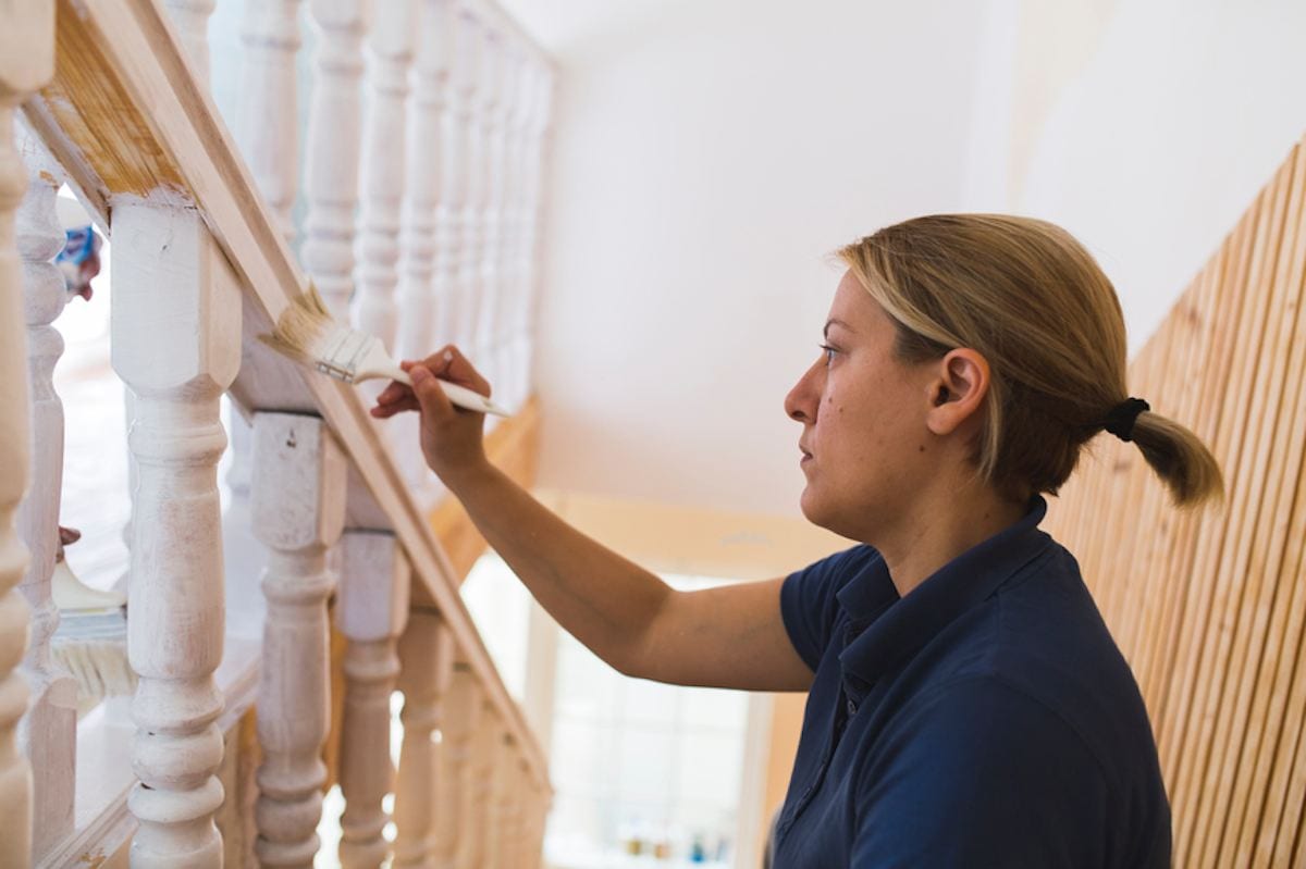 A blonde woman works on refinishing a banister.