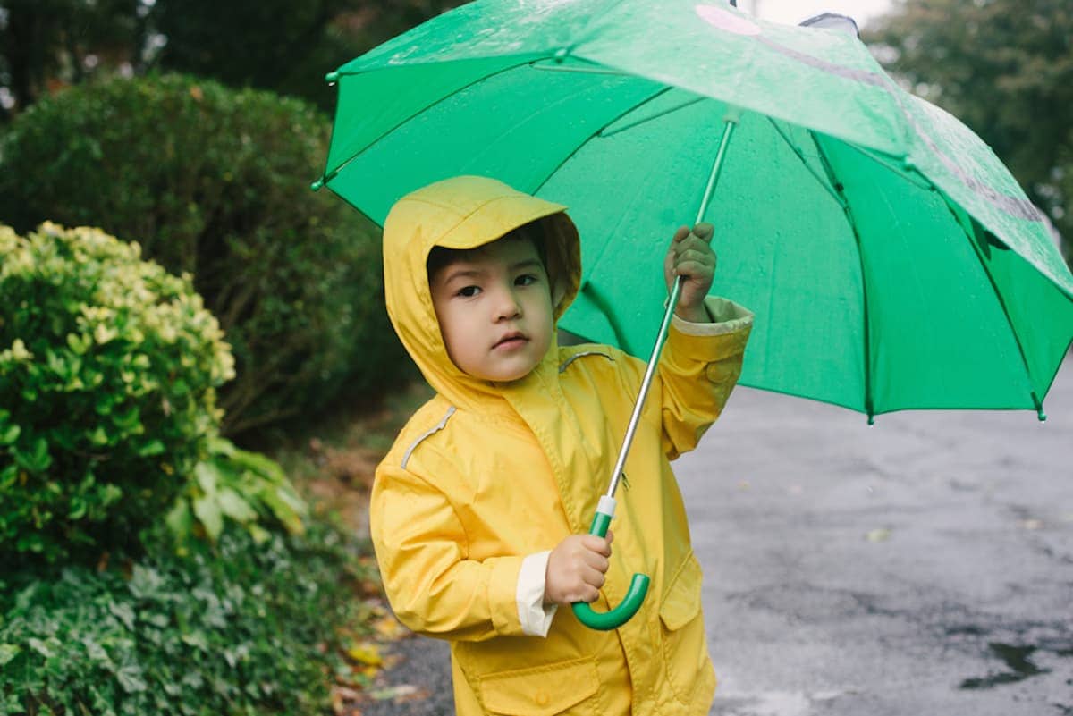 A little boy in a yellow raincoat holds a large green umbrella.