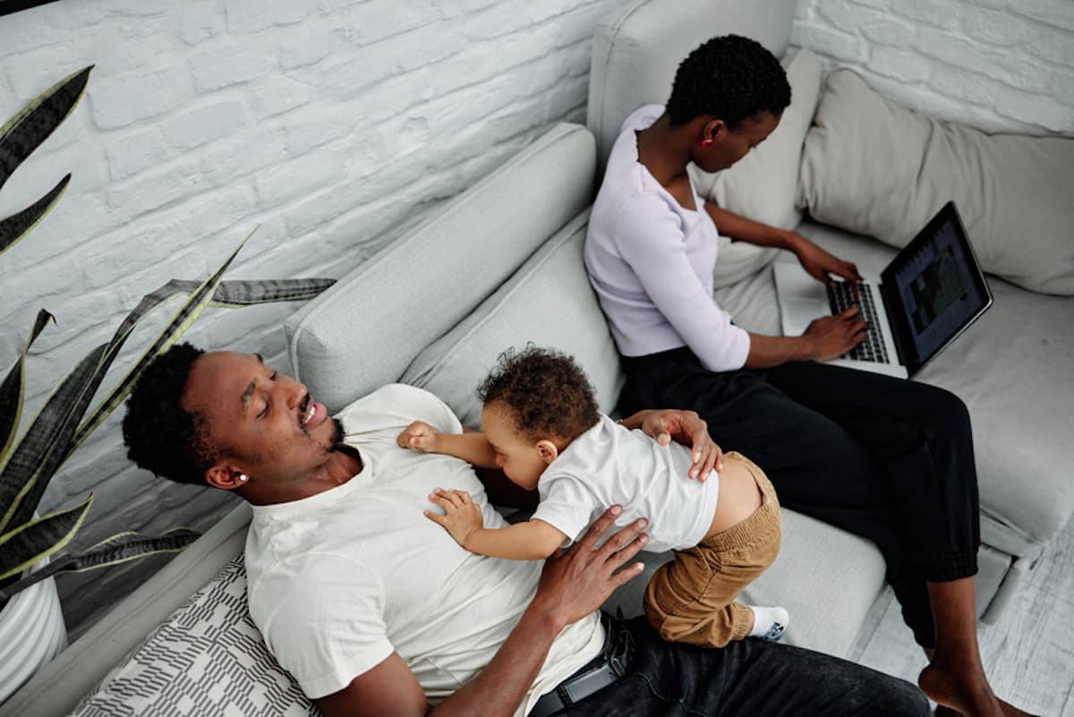 A Black man seated on a couch holds his infant while his wife works on a laptop next to him.
