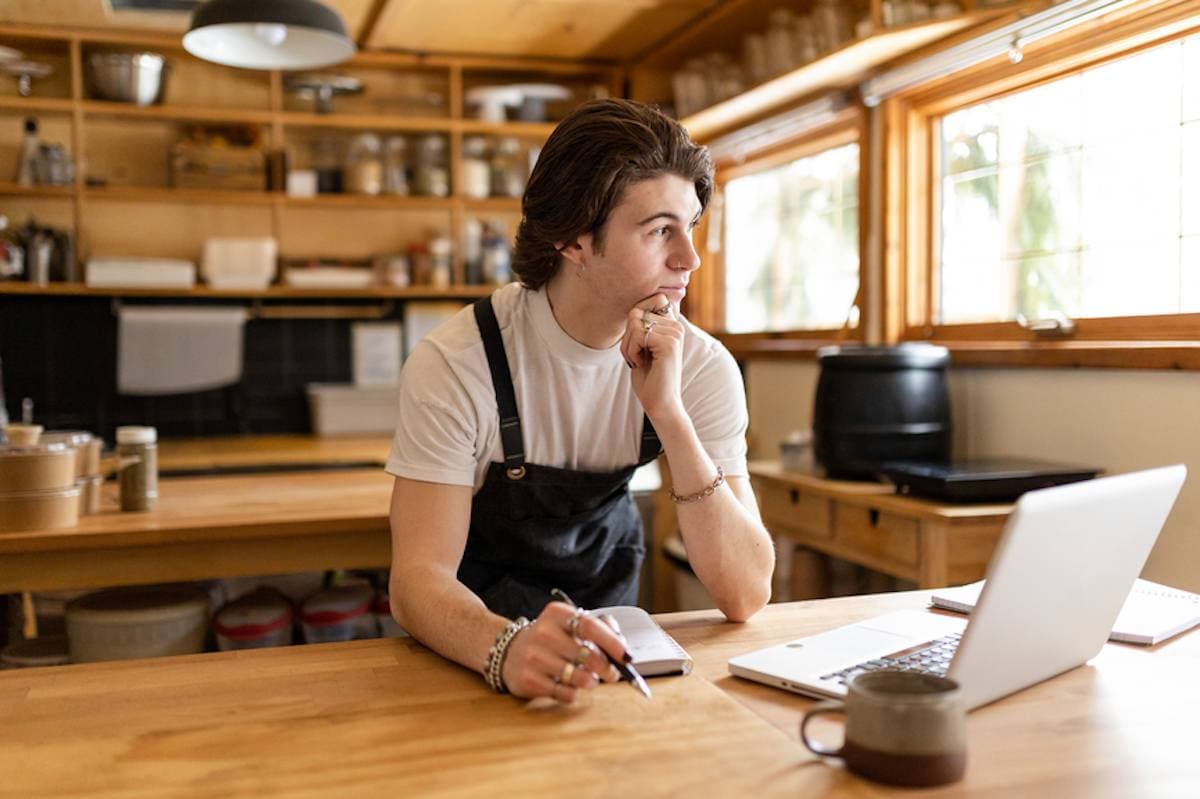 A male café employee with laptop thinks while looking out a window