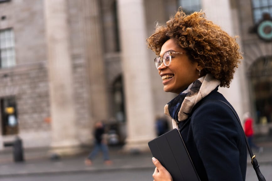 A Black woman smiles as she walks down the street holding a laptop case.