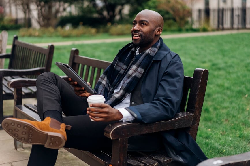 A Black businessman holding a tablet and a coffee relaxes on a park bench.