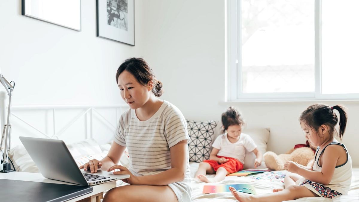 young mom working on a laptop with small children playing behind her