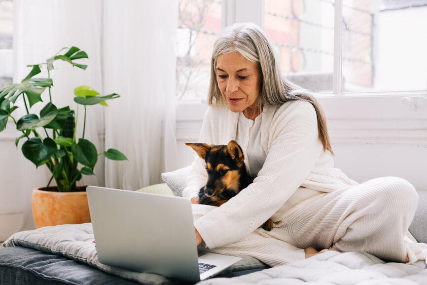 A silver haired woman with a dog in her lap types on a laptop.