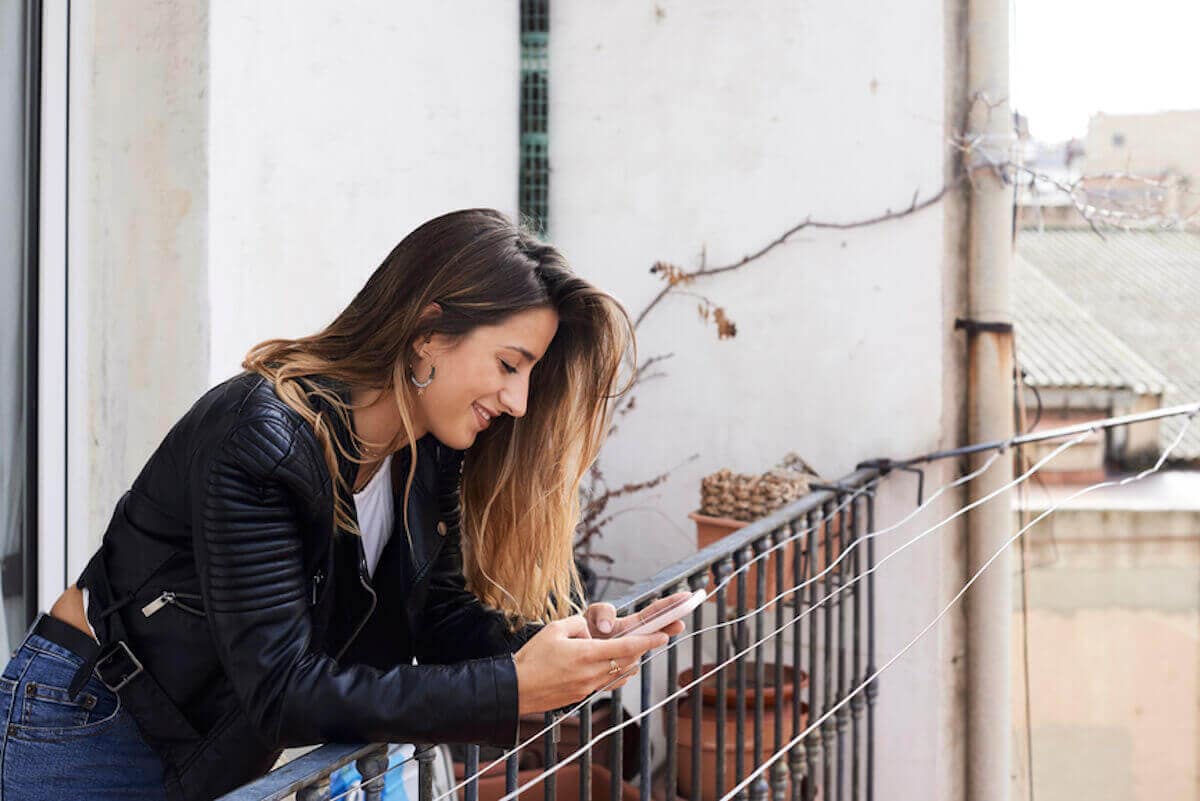 Smiling young woman leans on balcony at home and uses smartphone