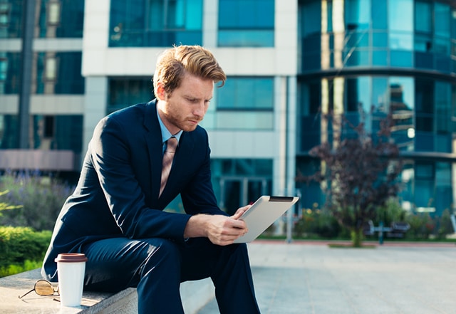 young man in business attire reading on his tablet outside of an office building