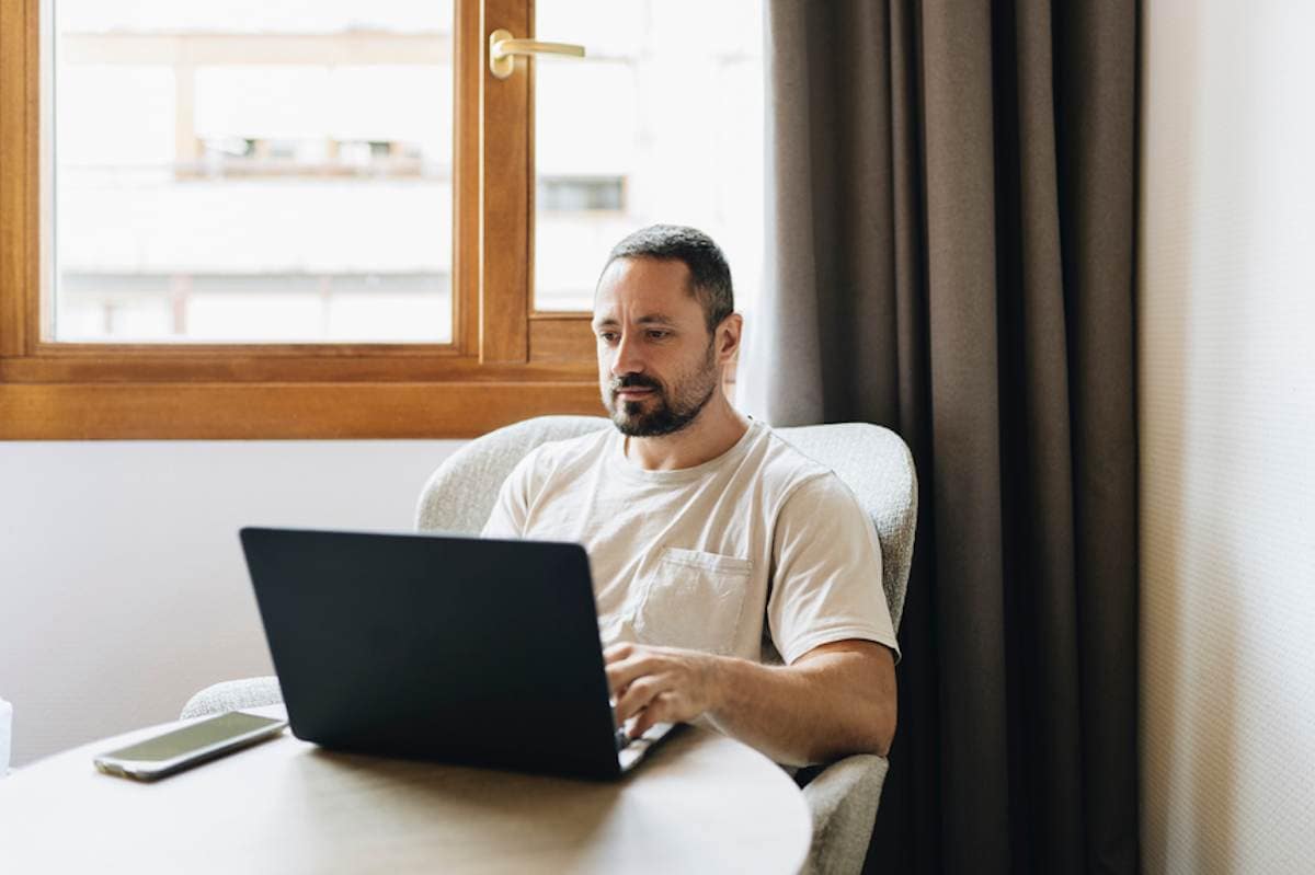 Bearded man in tee shirt focuses on laptop while sitting in chair at home