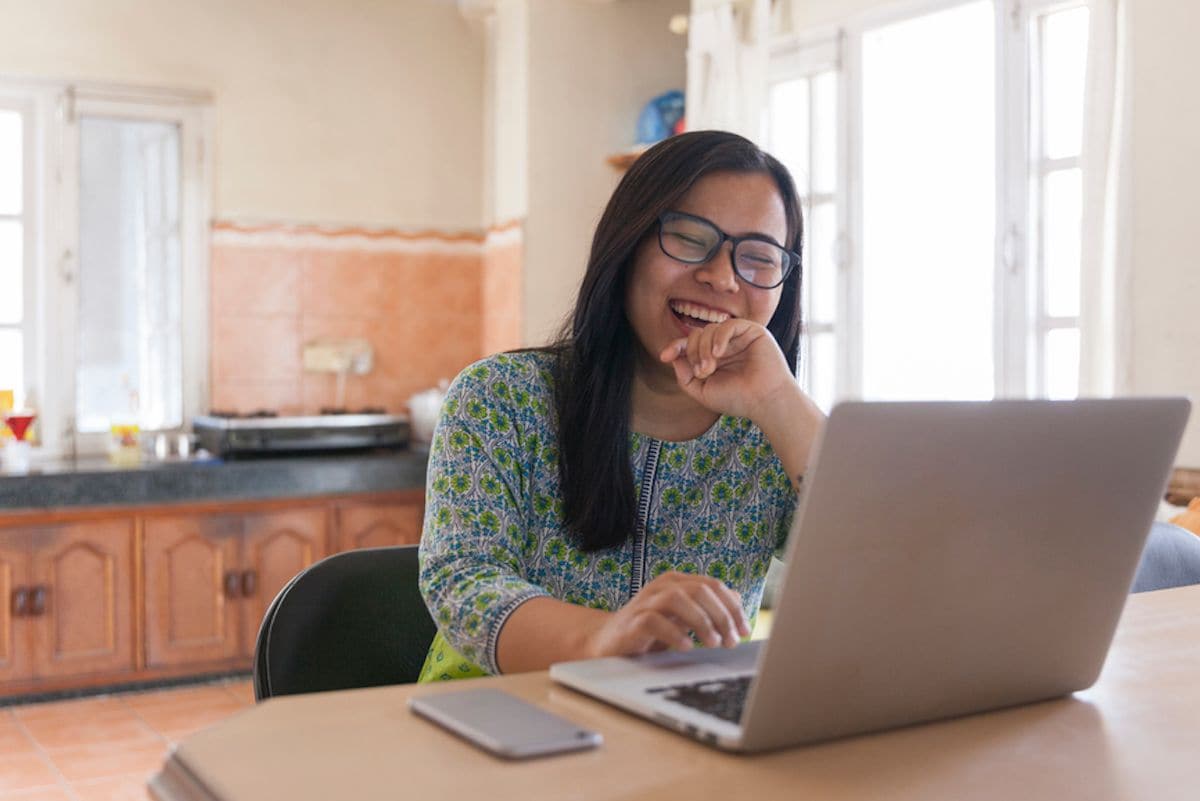 A woman seated at her laptop looks at the screen and smiles.