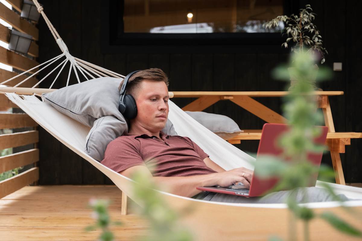 Male freelancer wearing headphones uses a laptop while lying in hammock