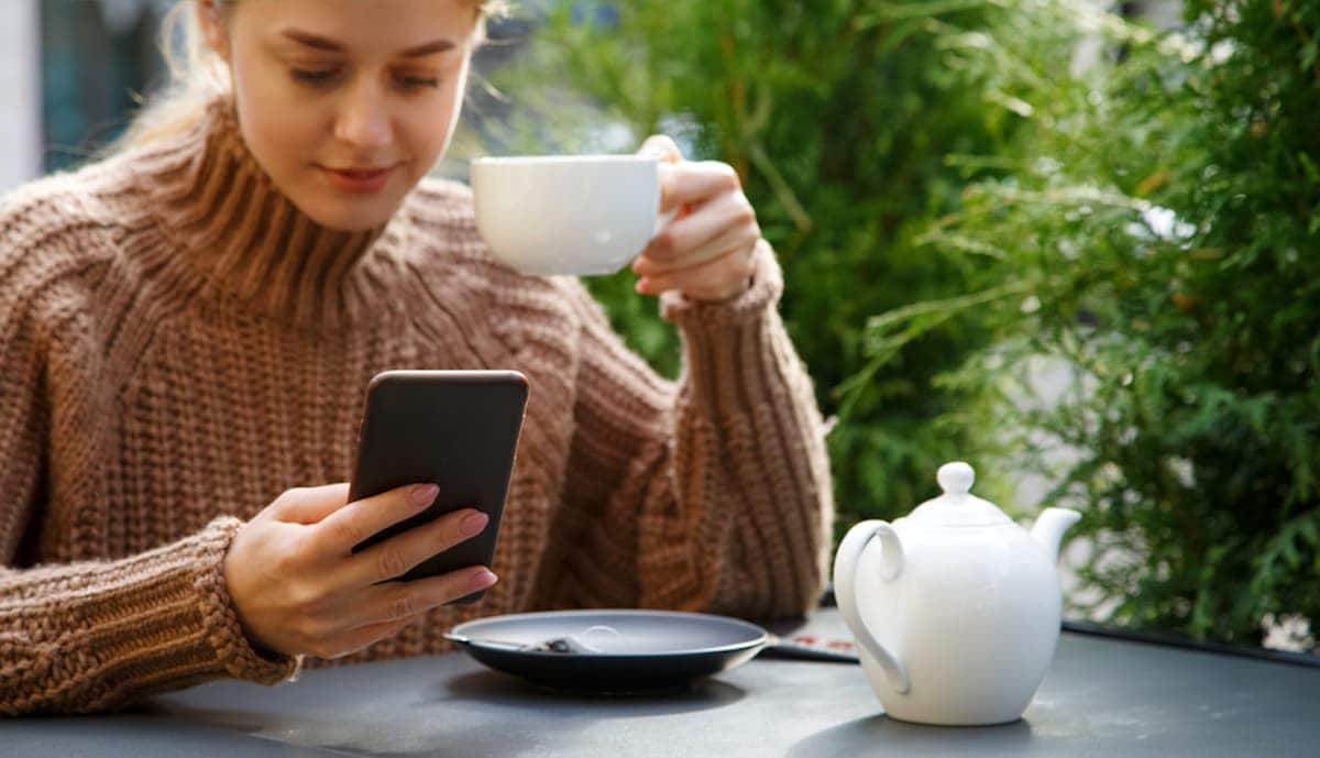A woman holding a cup of coffee focuses on her phone screen.