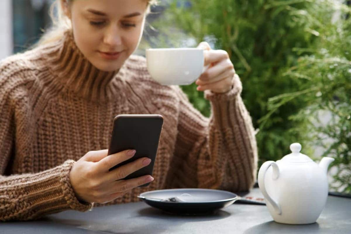 A woman holding a cup of coffee focuses on her phone screen.