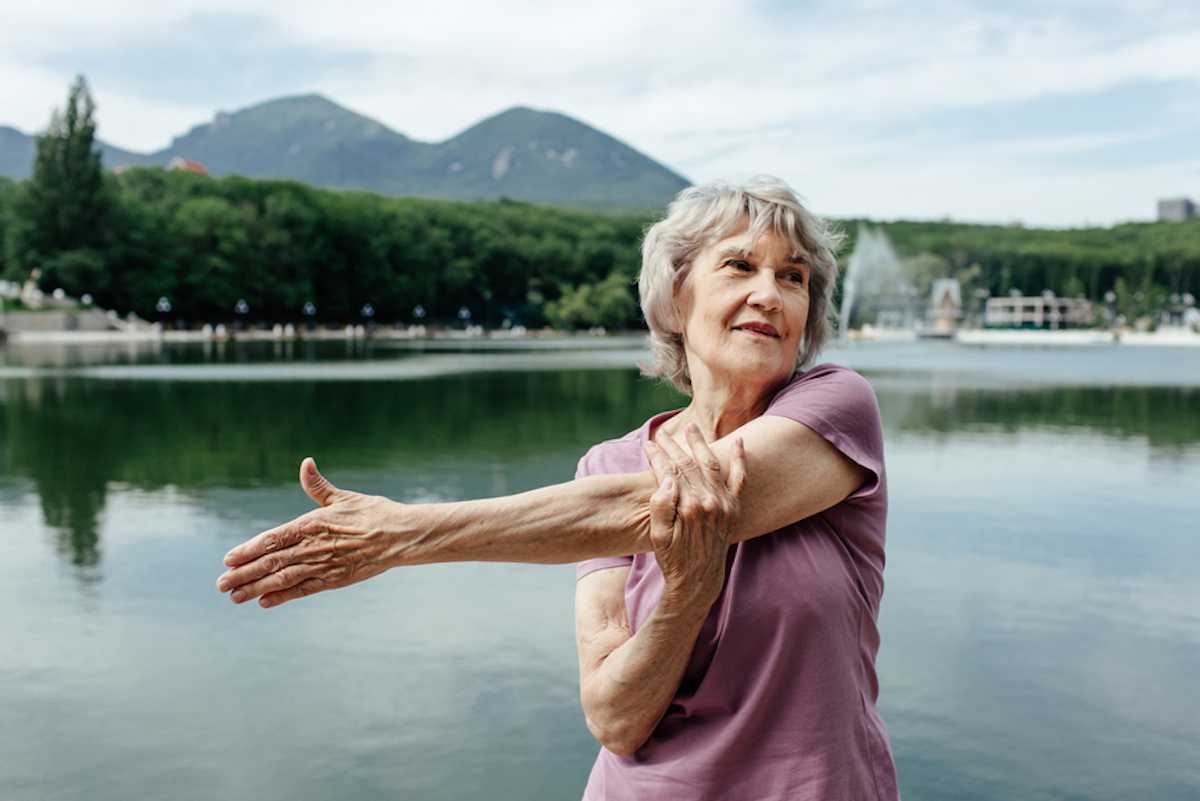 Senior woman in activewear does arm stretch by lake near mountains