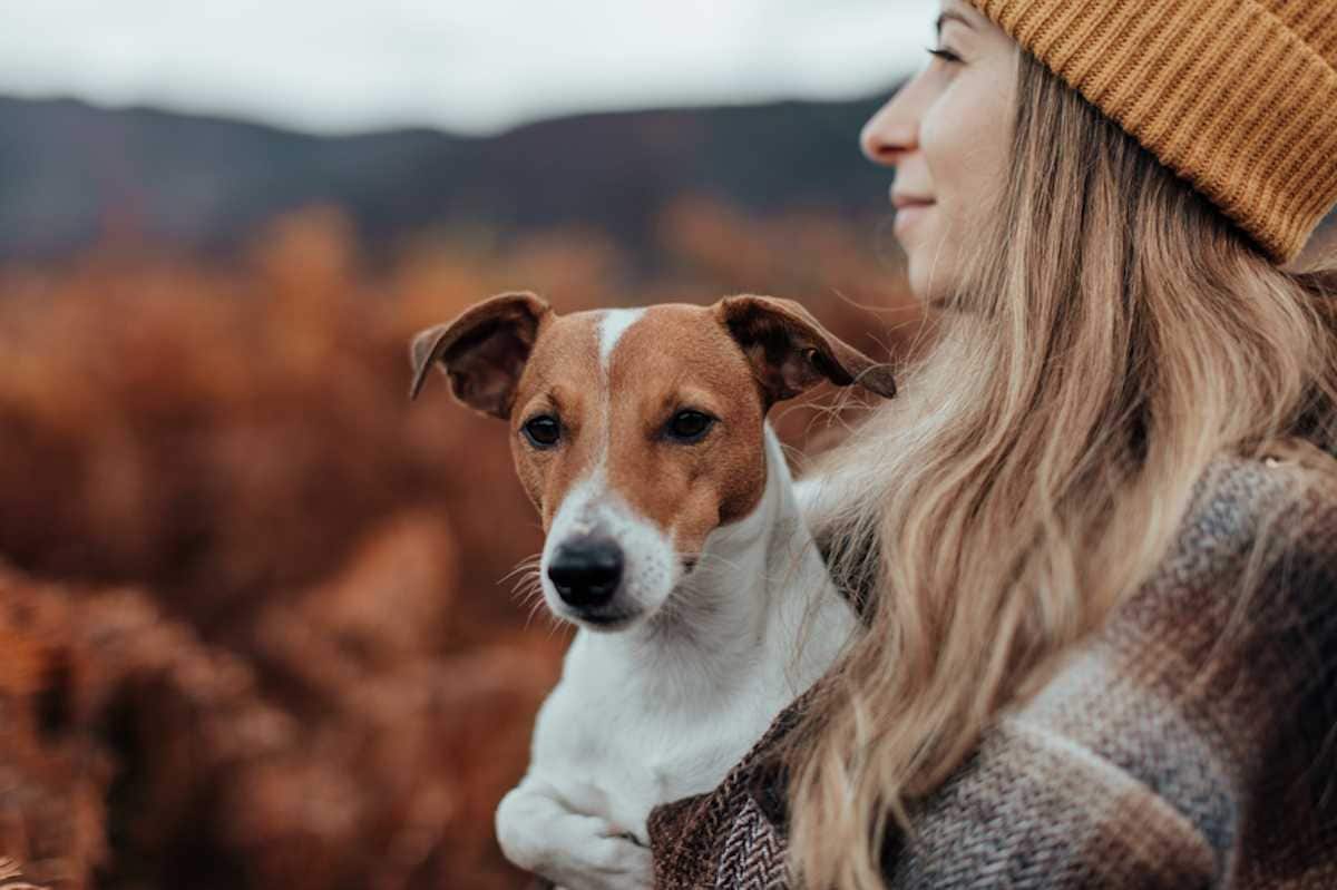 Young woman in stocking cap holds her dog and looks at fall landscape