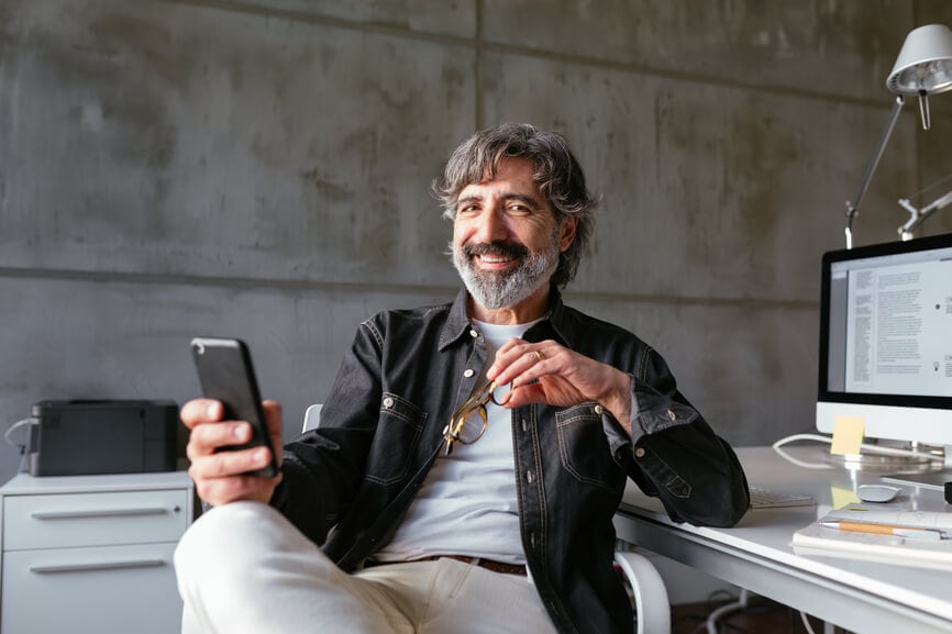 Delighted bearded senior employee with glasses smiling and looking at camera while sitting near desk and using smartphone during break in office.