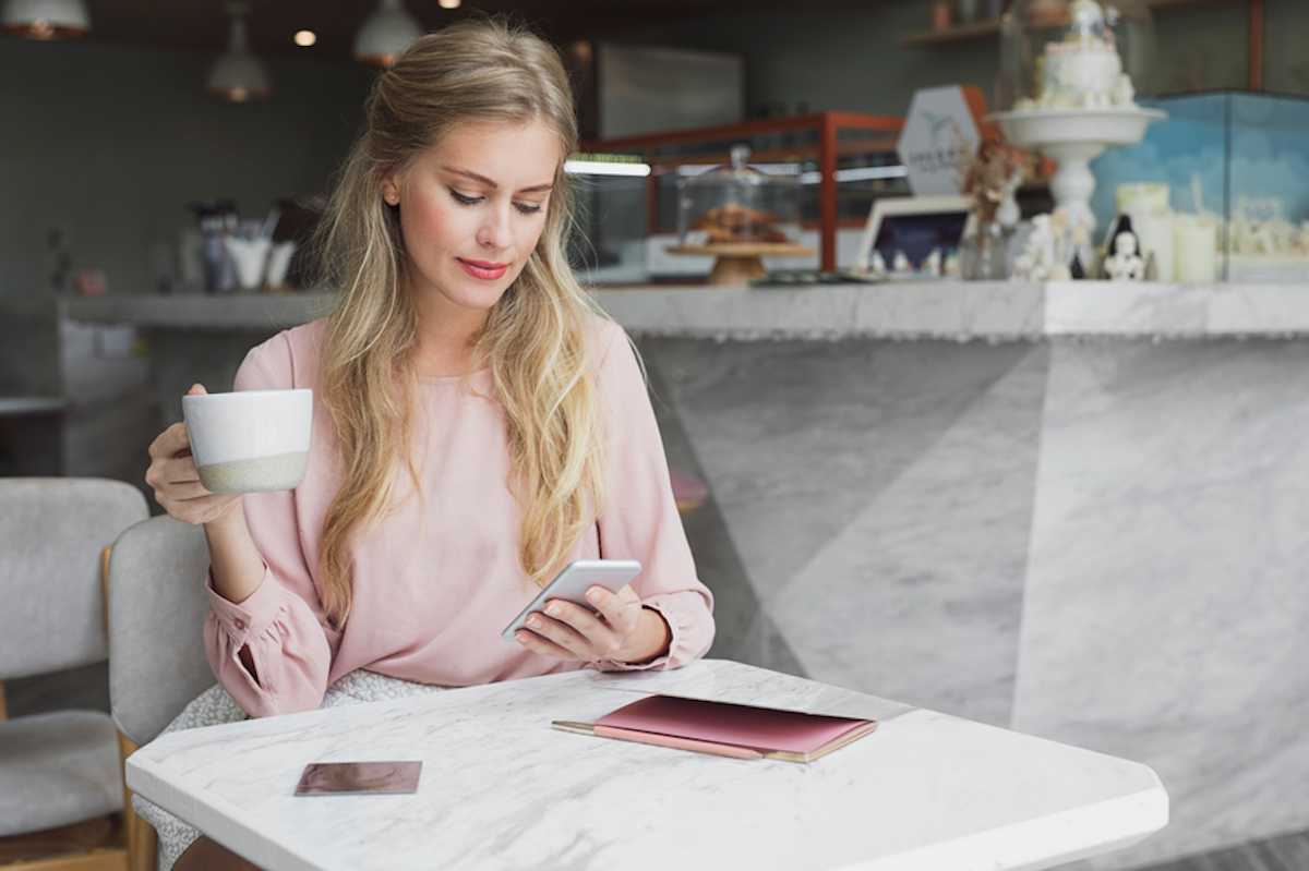 Young woman sitting alone at coffee shop holds mug of coffee and checks her smartphone